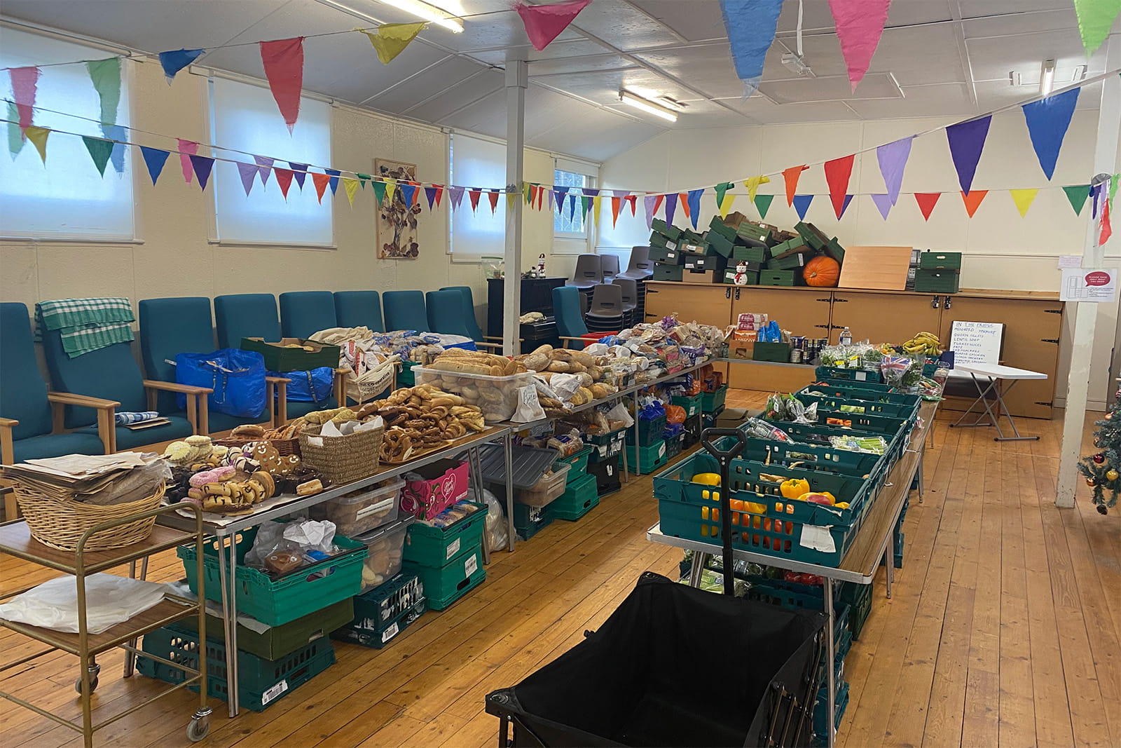 Wide view inside a community hall set up for a community fridge session, with long tables and crates of surplus bread, groceries, and fruit and vegetables under colourful bunting.