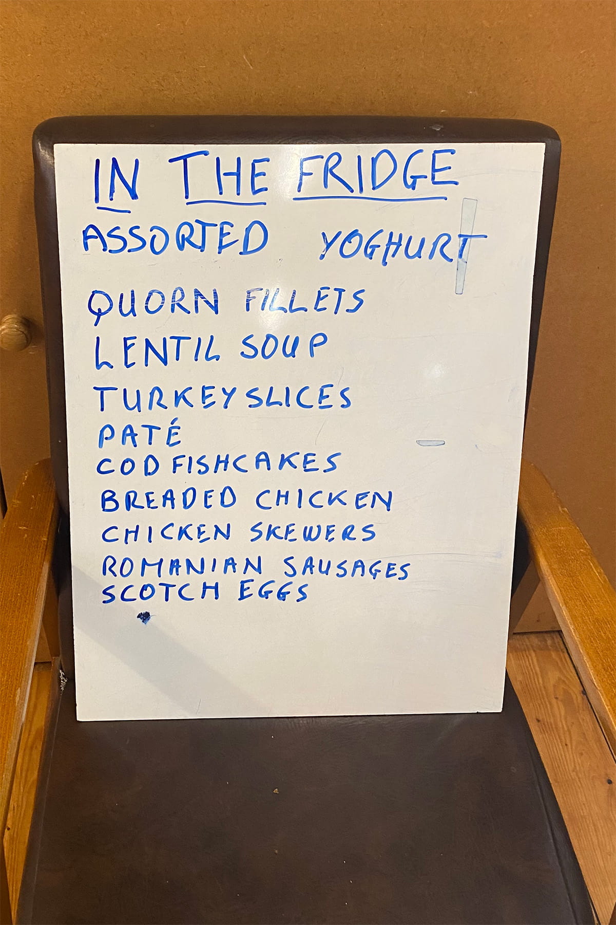 Whiteboard sign titled “IN THE FRIDGE” listing available items: assorted yoghurt, Quorn fillets, lentil soup, turkey slices, pâté, cod fishcakes, breaded chicken, chicken skewers, Romanian sausages, and Scotch eggs.