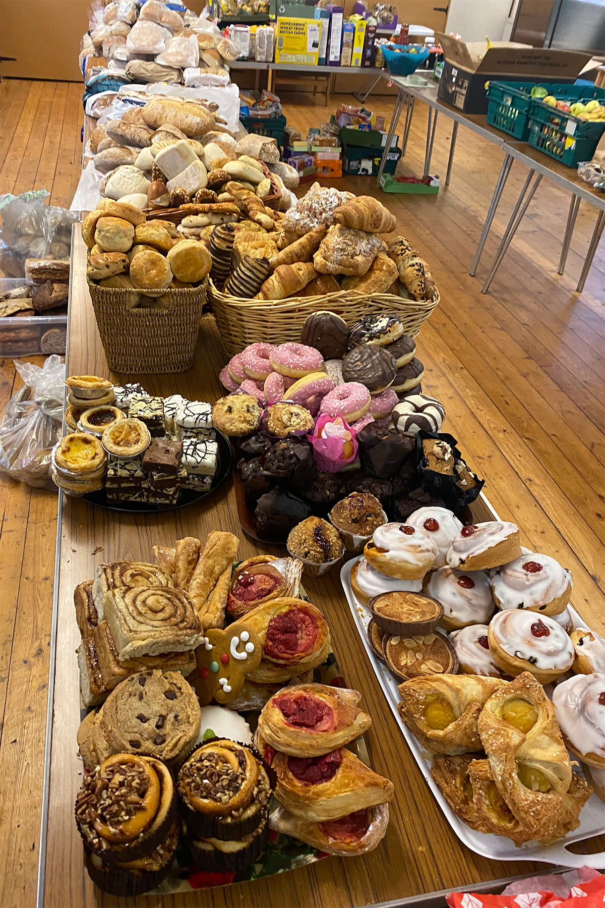 Close-up of a community fridge distribution table piled with surplus baked goods—breads, rolls, pastries, donuts, muffins, and iced buns—laid out in baskets and trays in a hall.