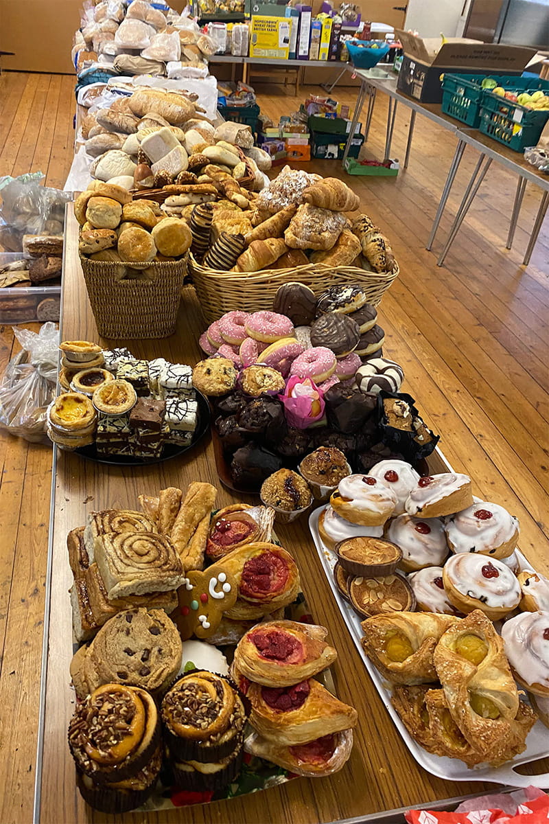 Close-up of a community fridge distribution table piled with surplus baked goods—breads, rolls, pastries, donuts, muffins, and iced buns—laid out in baskets and trays in a hall.