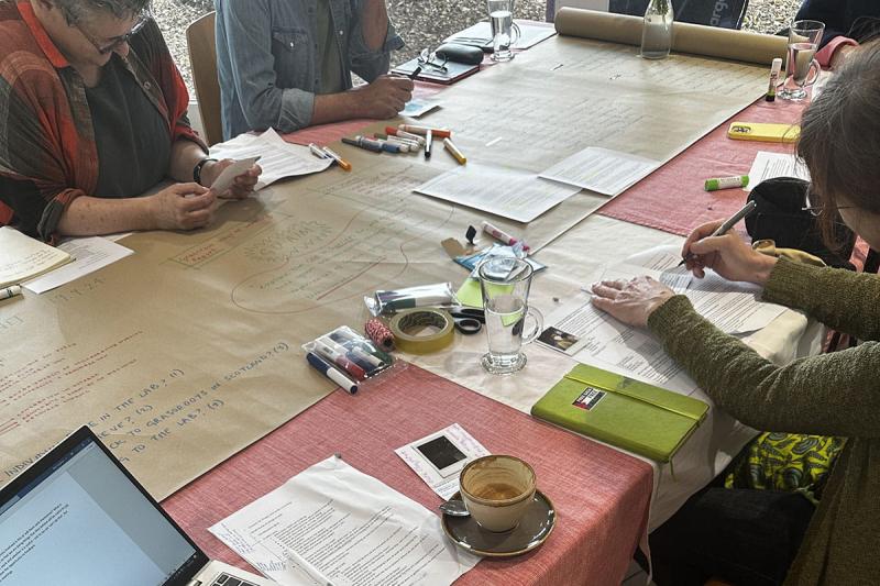 Individuals seated on both sides of a long table covered with a red cloth, featuring a long strip of brown paper with scattered pens, papers, coffee cups, and a laptop.