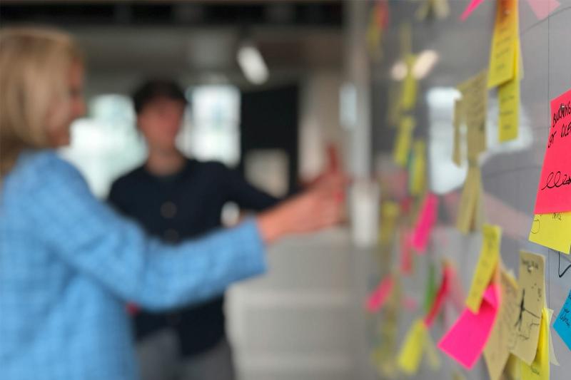 Two people gesture towards a whiteboard filled with various sticky notes during a mind-mapping session.