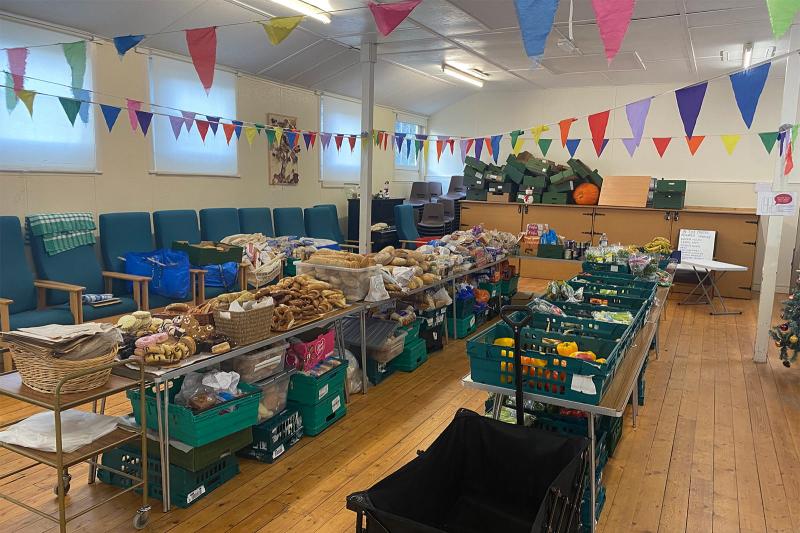 Wide view inside a community hall set up for a community fridge session, with long tables and crates of surplus bread, groceries, and fruit and vegetables under colourful bunting.