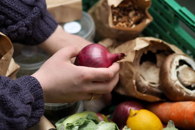 Close-up of someone selecting a red onion from a crate of surplus vegetables, including mushrooms, carrots and citrus fruit. 